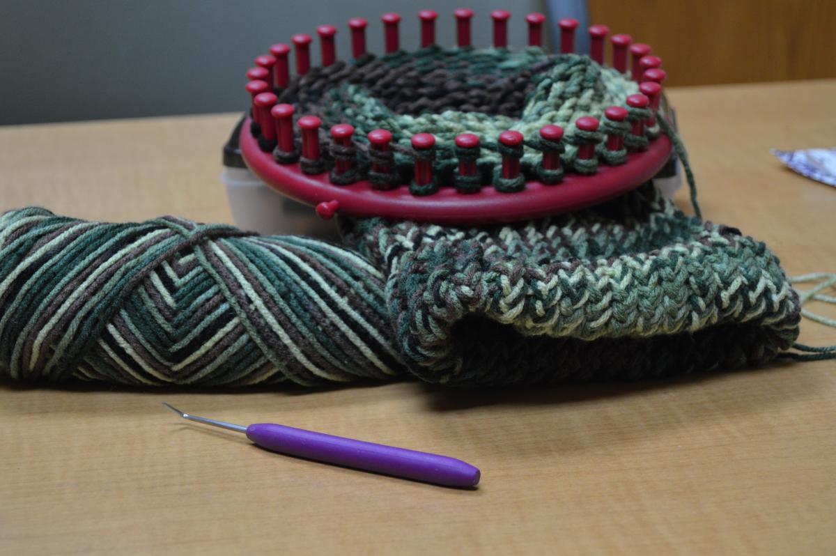 picture of a dark red round loom with camouflage colored yarn being knitted into a hat. A round loom tool is in the forefront.