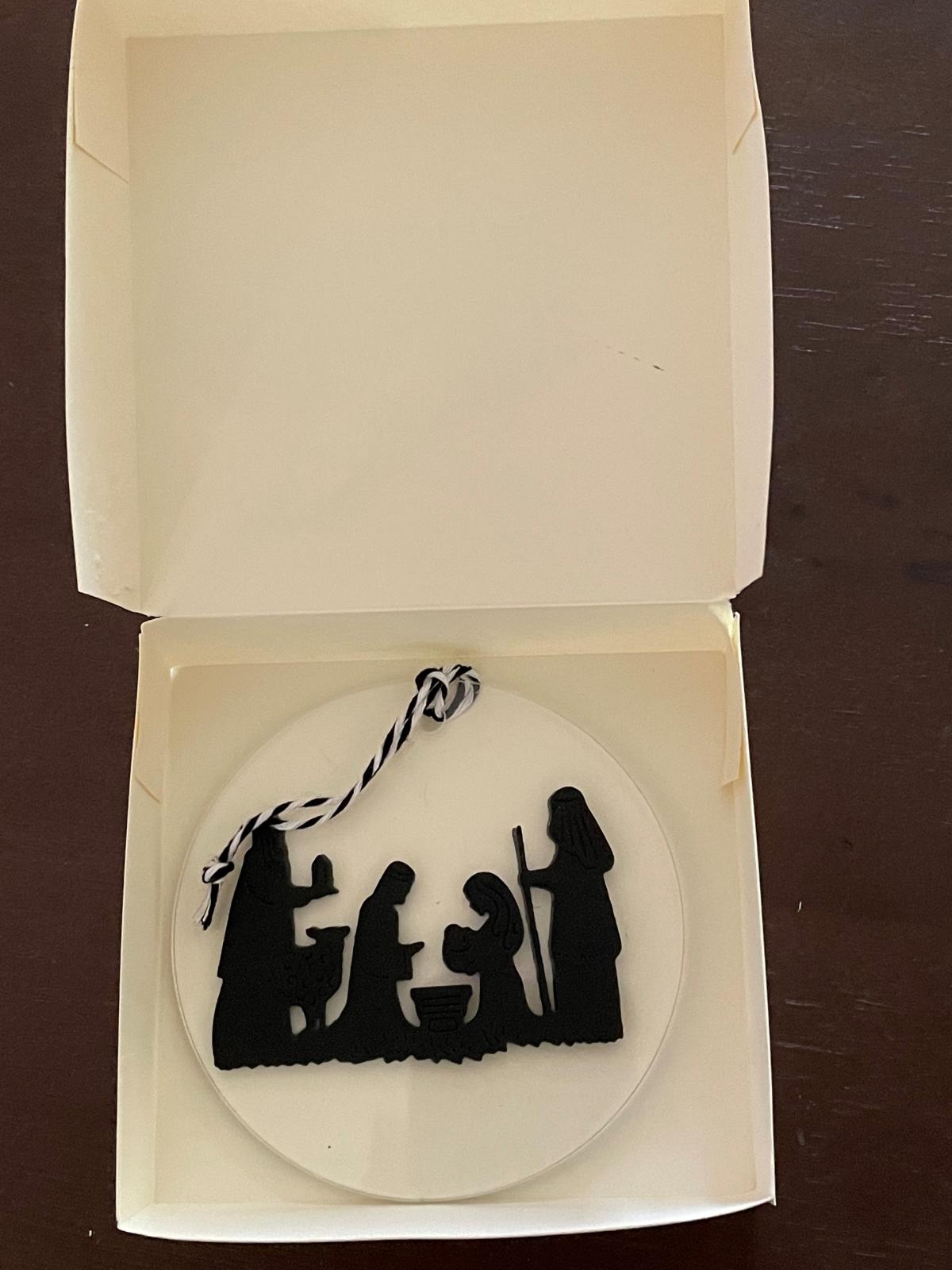 A white paper box containing an acrylic ornament with black and white string and a black nativity silhouette.